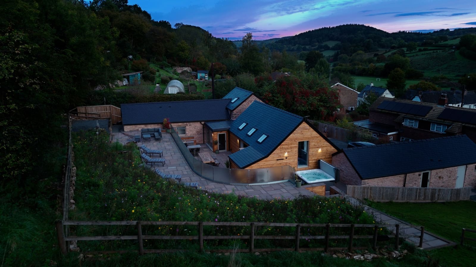 The Walnut Barn from the air at dusk, lit terraces and hot tub on a wooded hillside above the valley.