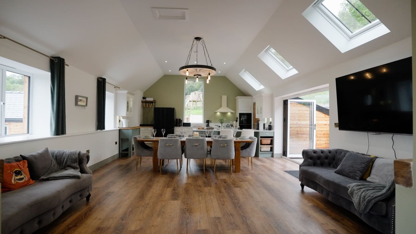 The vaulted living room: two velvet sofas facing the long dining table and kitchen, under roof lights and a cartwheel chandelier.