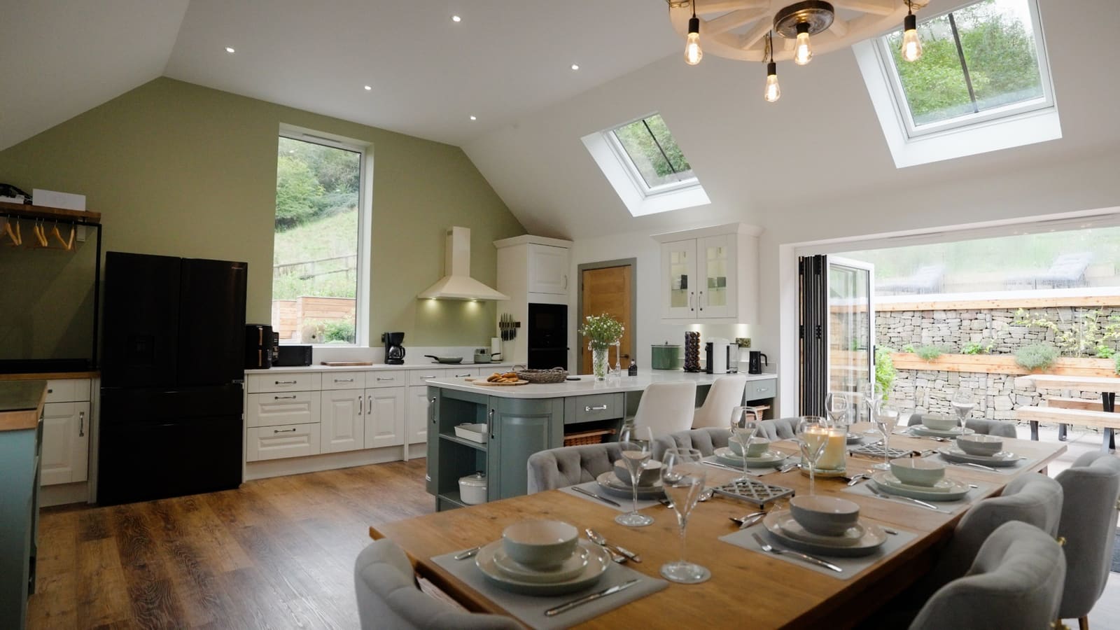The dining table laid for a meal, with the kitchen, tall window and roof lights beyond.