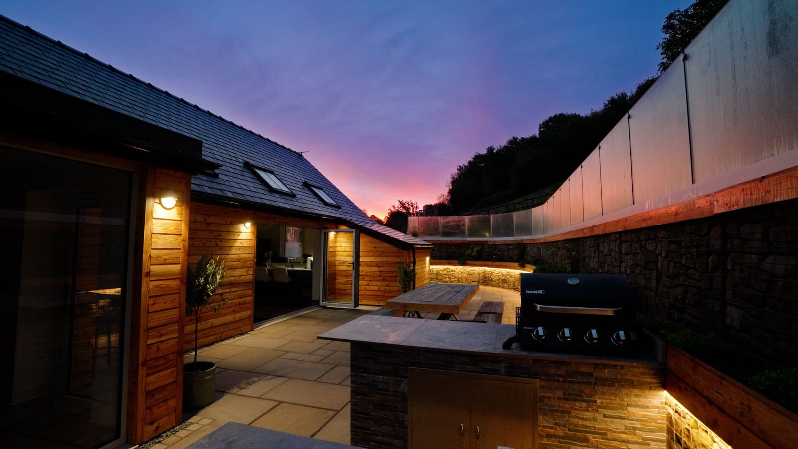 The courtyard at sunset: lit timber walls, the outdoor kitchen and the long table.