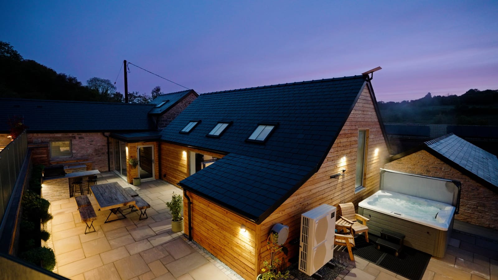 The barn at dusk, its wall lights on, with the courtyard table and the private hot tub.
