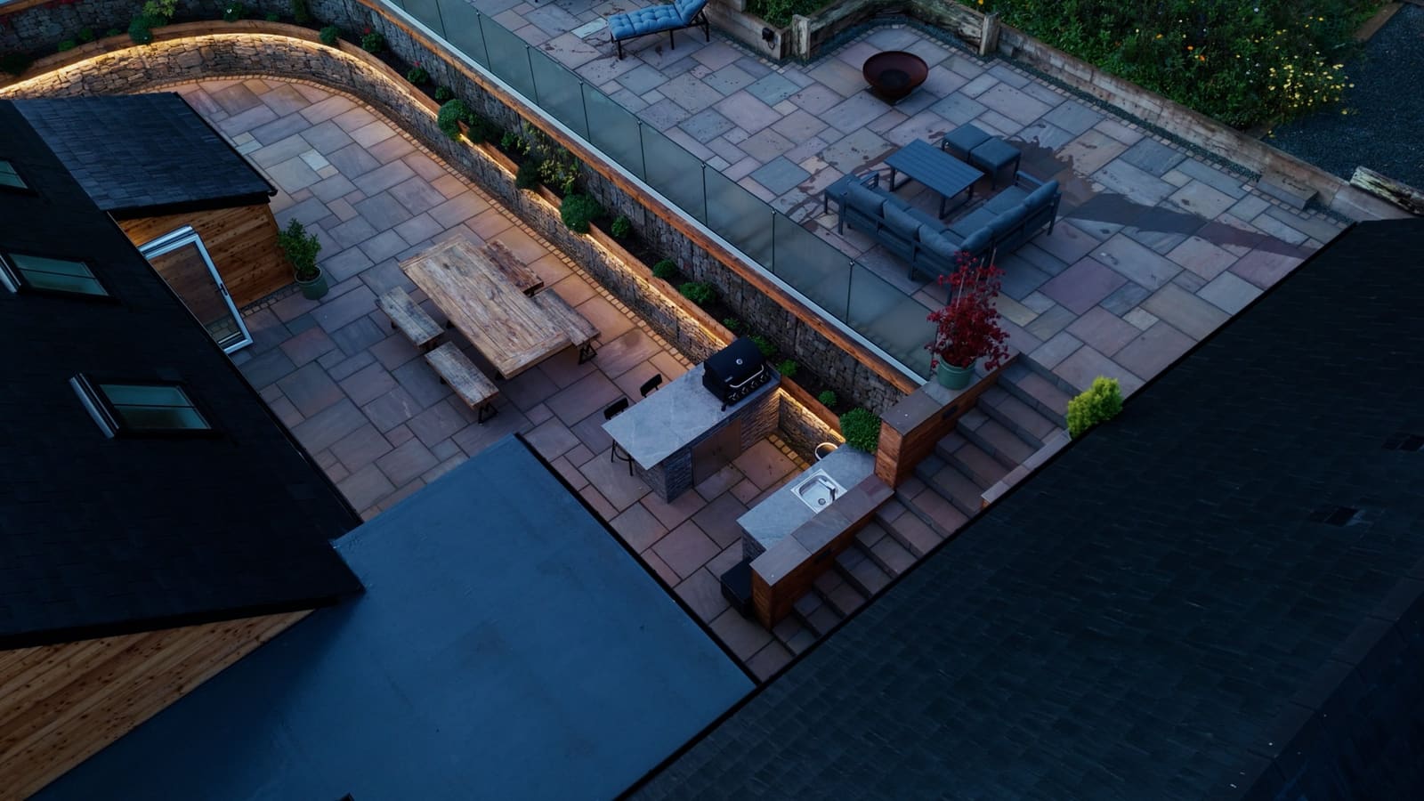 The terraces from above at dusk: the lit stone wall, outdoor kitchen, dining table and upper lounge.
