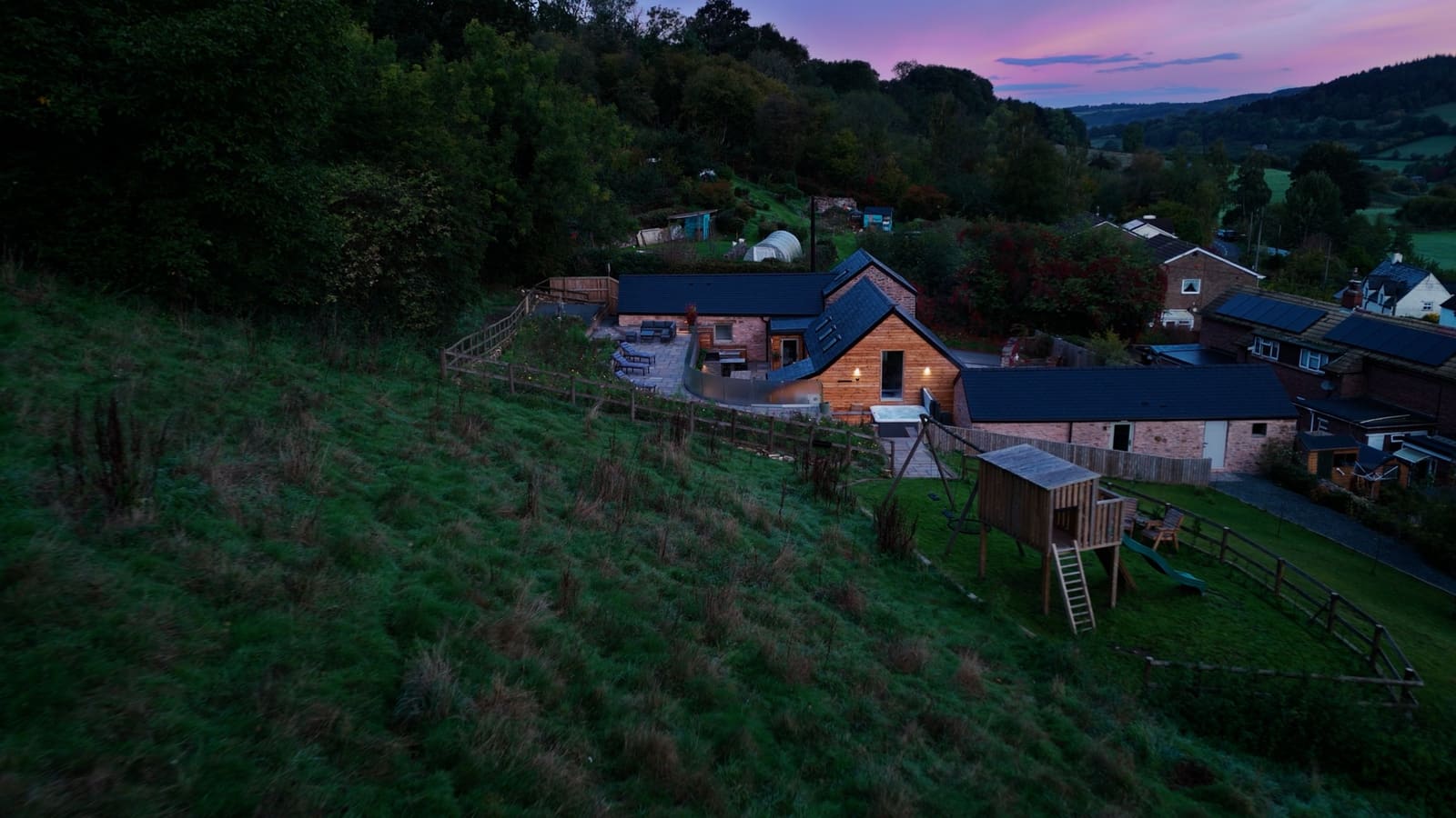 The barn from the hillside at dusk, with the play area, the village and the valley under a pink sky.