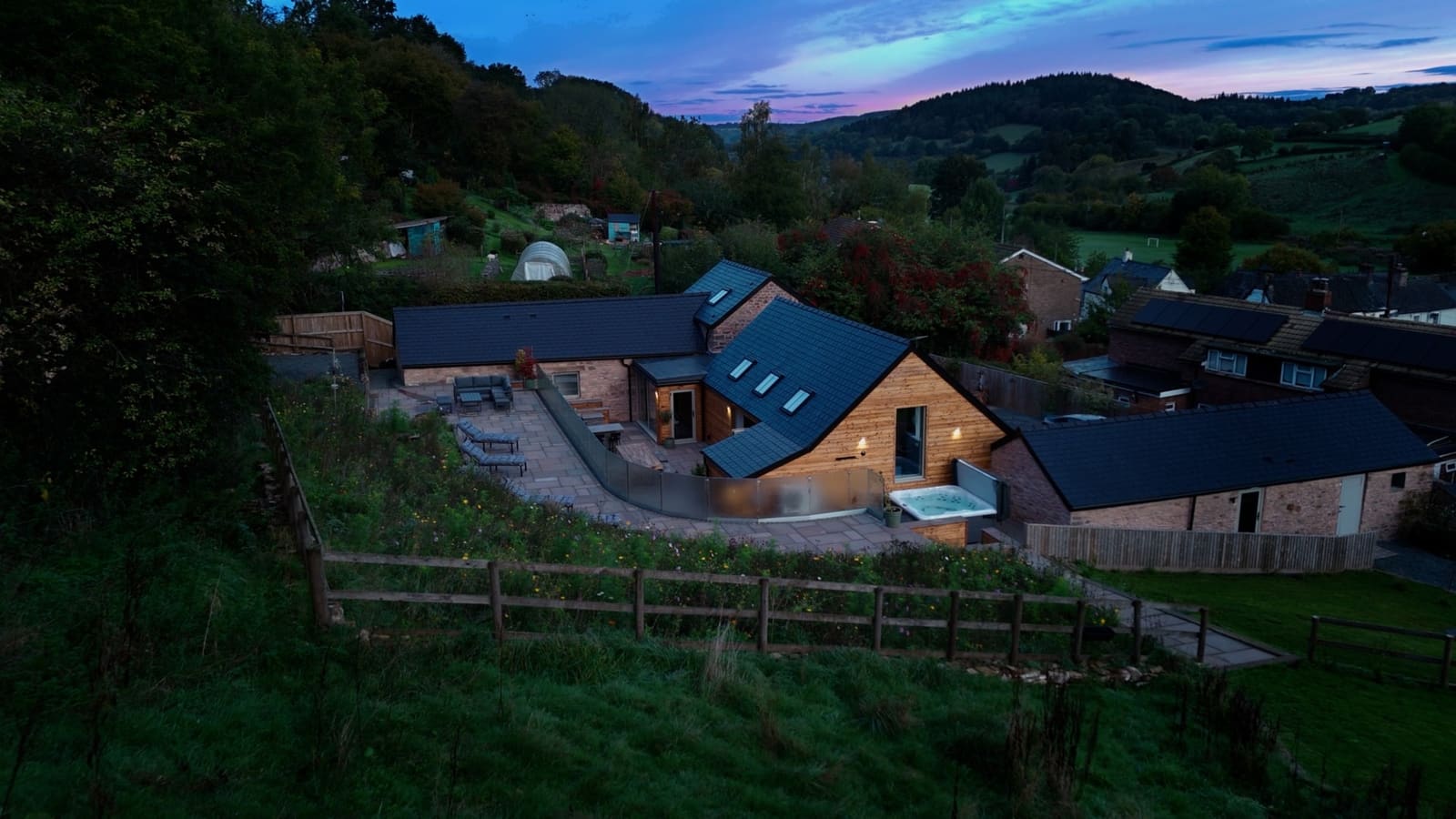 The Walnut Barn from the air at dusk, its terraces and hot tub lit, with the village and wooded hills beyond.