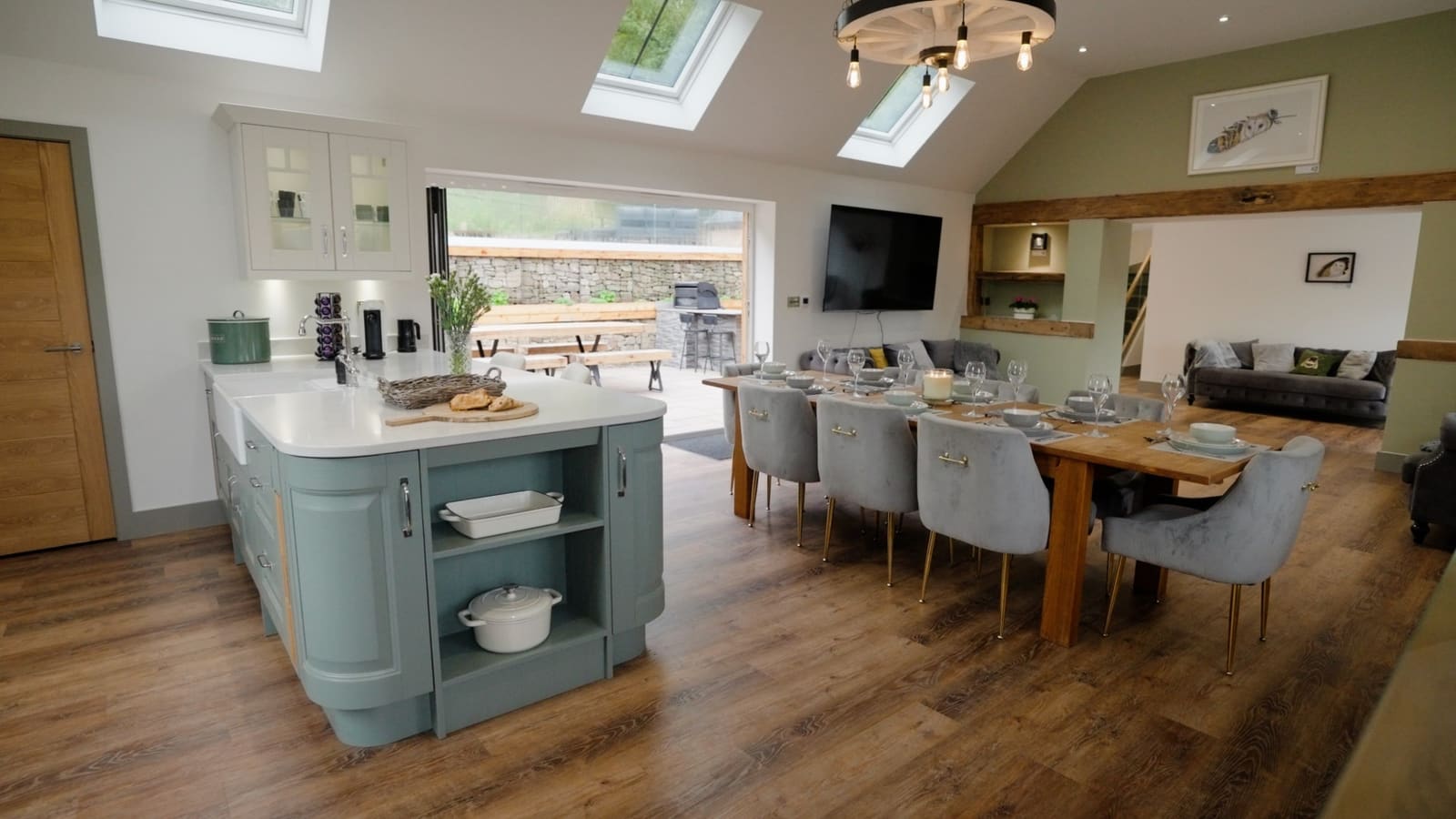 The open-plan kitchen and dining room: a sage-green island, a long oak table and roof lights in the vaulted ceiling.